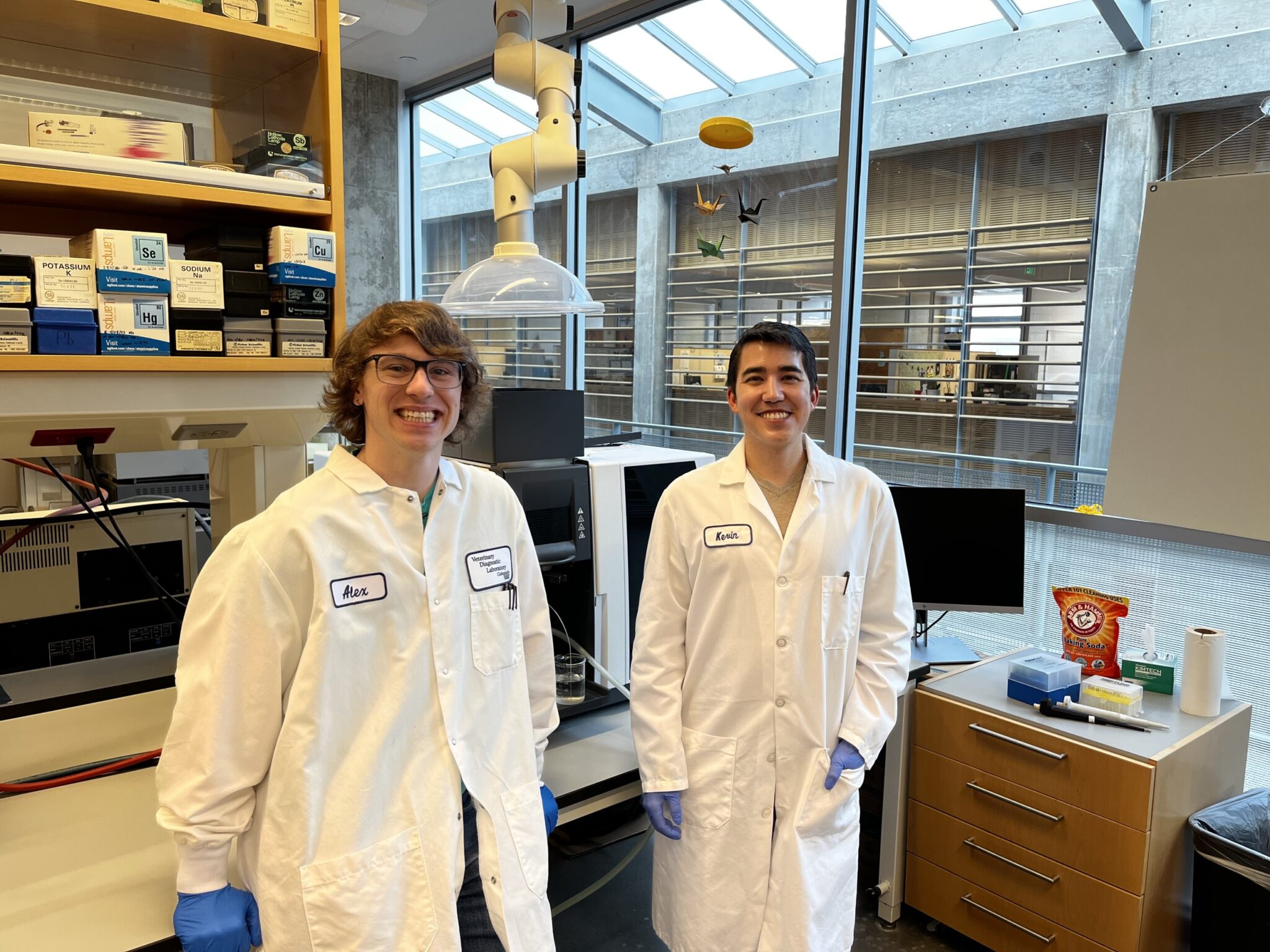 Two individuals wearing white laboratory coats and protective gloves stand inside a research laboratory. Behind them is a large window with a view of an outdoor area featuring concrete structures and metal railings. The laboratory contains equipment such as shelving with boxed supplies, a microscope, a fume extraction hood, and various instruments on countertops.