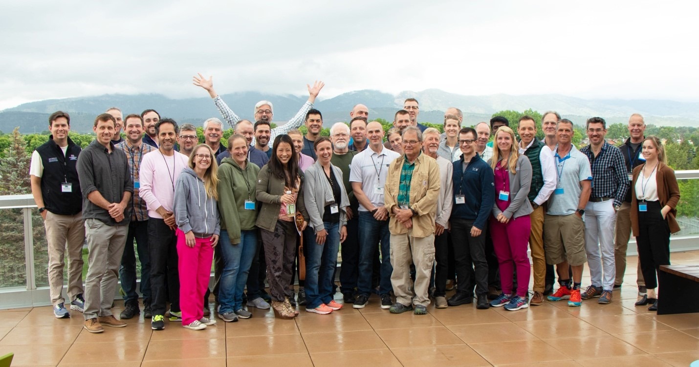 A large group of people stand closely together on an outdoor terrace with mountains and cloudy skies in the background. The group is arranged in several rows, wearing a mix of casual and business‑casual clothing, with many individuals wearing name badges. One person near the back raises both arms enthusiastically above their head. The terrace has a smooth tiled surface, and greenery surrounds the area beyond the railing.