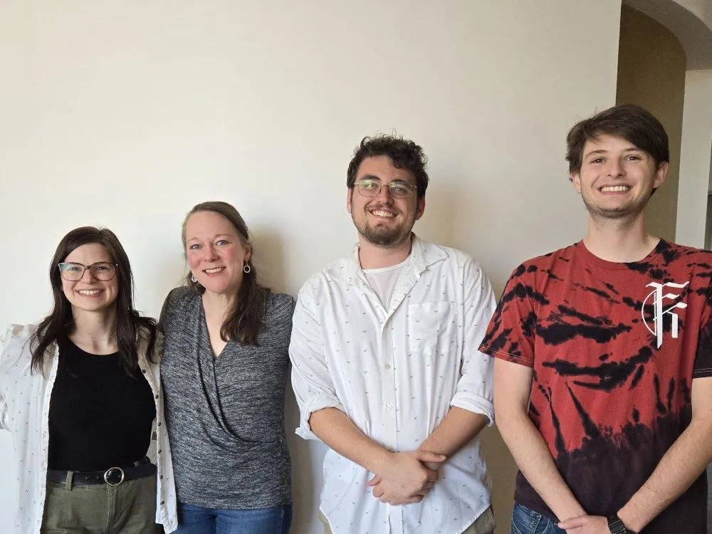 Four individuals stand side by side indoors against a plain light-colored wall. They are wearing casual clothing, including button-up shirts, a patterned top, a graphic T-shirt, and belted pants. They are positioned close together, facing forward, with soft natural light illuminating the scene.
