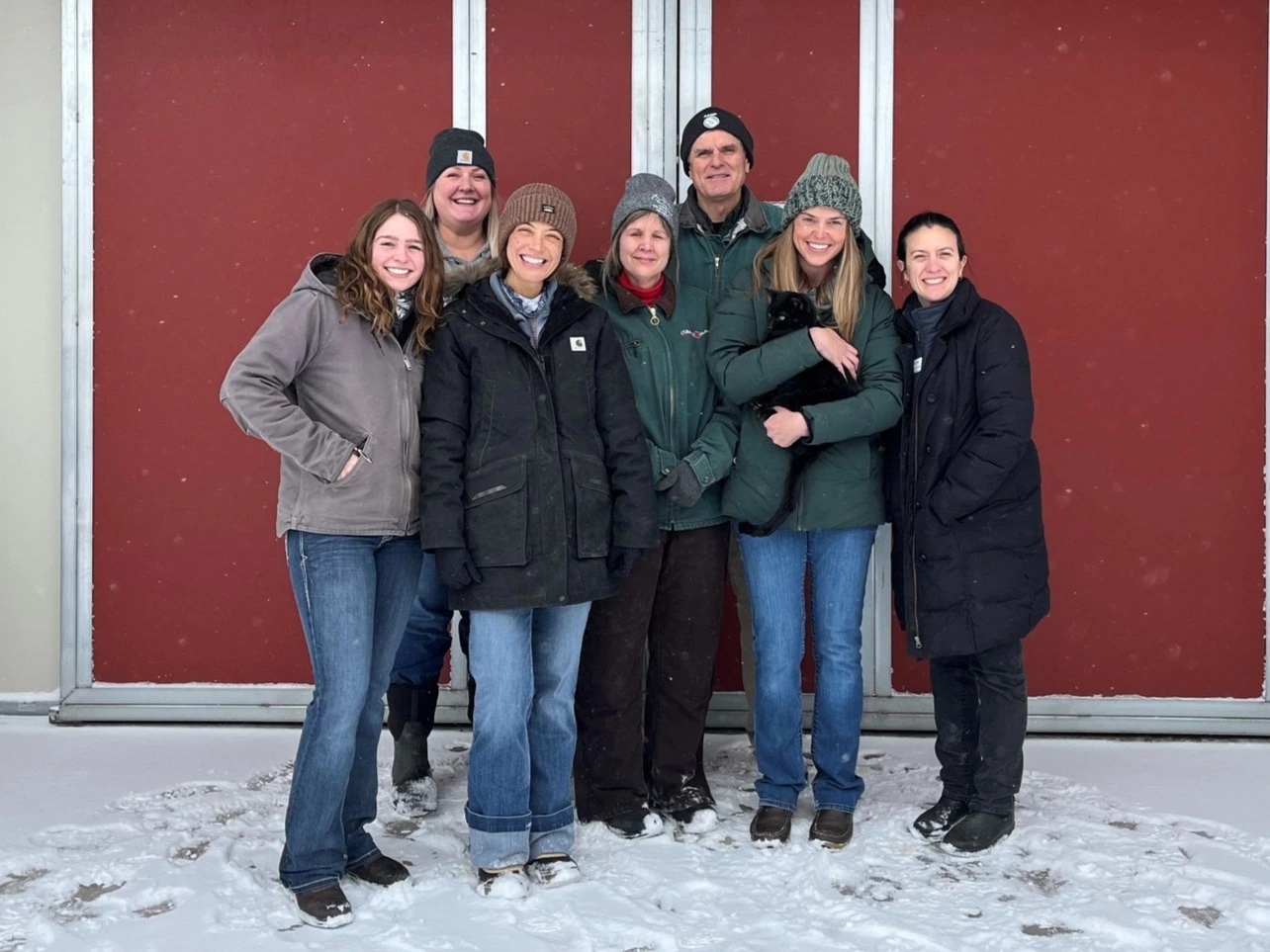 A group of eight people stand together outdoors in front of large red metal doors. They are dressed in winter clothing such as heavy coats, hats, gloves, and boots, and snow is visible on the ground around them. One person near the right side of the group is holding a small black animal wrapped in their arms. The scene appears cold, with light snow on the individuals’ clothing and footwear.