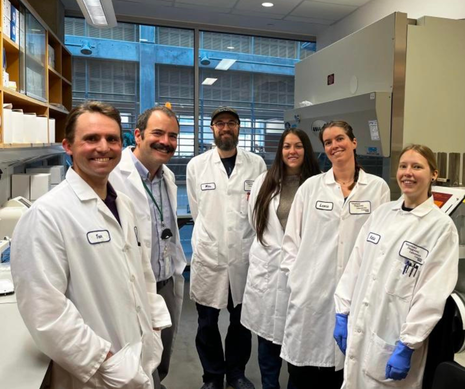 A group of six individuals wearing white laboratory coats stand together inside a research lab. Shelving with boxed supplies, a biosafety cabinet, microscopes, and various laboratory equipment surround them. Large windows in the background reveal an exterior building with a grid-like structure. Several members of the group wear gloves or ID badges, and they are positioned closely in the center of the lab workspace.