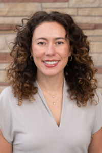 A woman smiles at the camera, with curly brown hair and a brick background