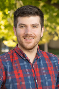 a man with dark short hair and short beard smiles at the camera, with a blue and red plaid shirt and green trees in the background