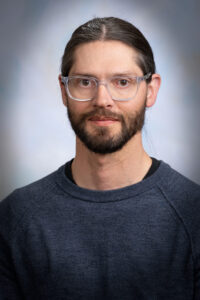 a man with dark long hair pulled back and a beard, looks at the camera, with a gray and white background
