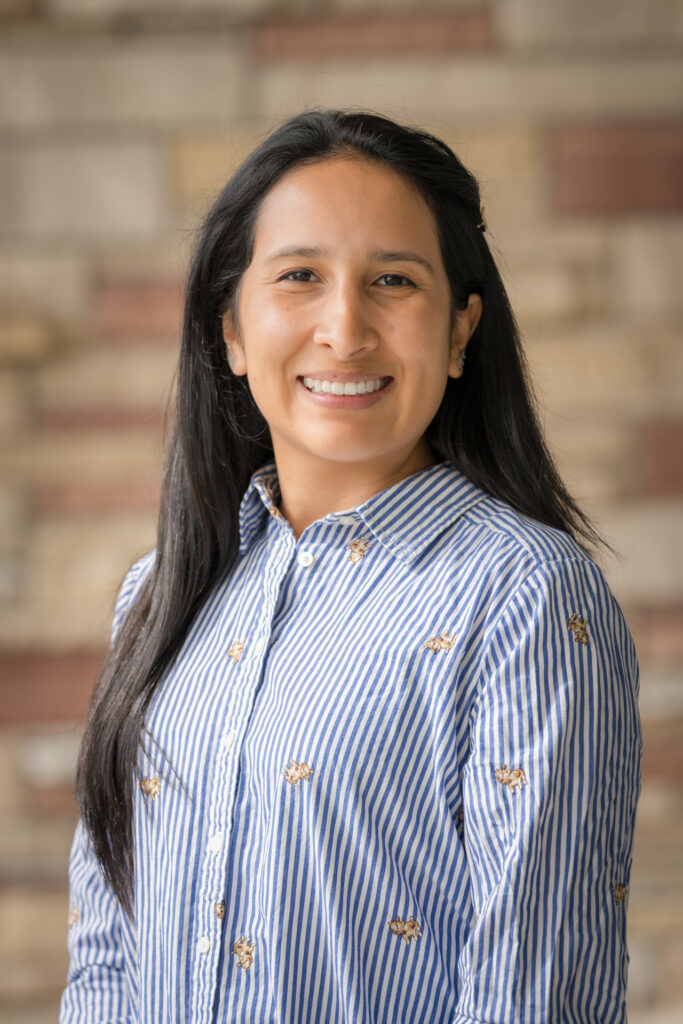 a person smiles at the camera, with long black hair and a red and white brick background