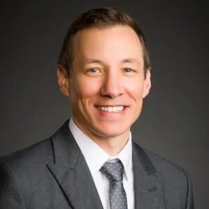 a man looks at the camera and smiles, wearing a gray suit with a gray background