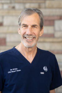 professional portrait of a gray-haired man with a short beard wearing dark blue scrubs against a brick wall background