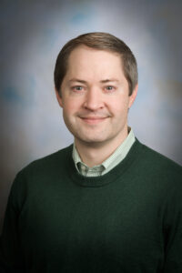 professional portrait of a smiling, brown-haired man wearing a green sweater