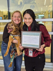 two people pose with a certificate and a little brown dog