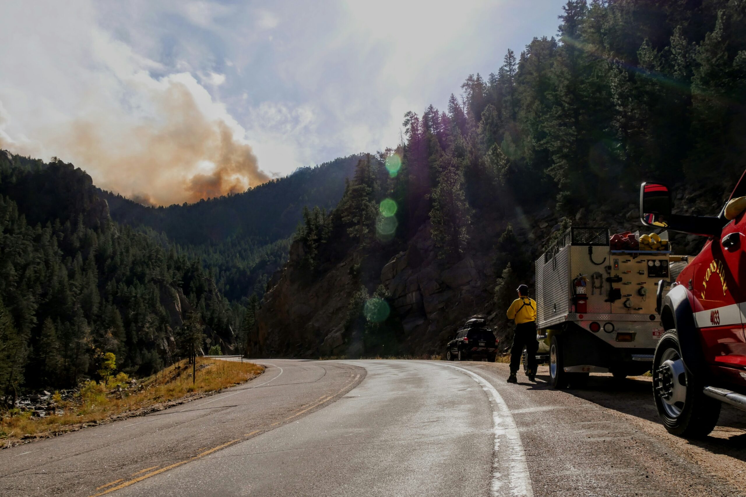a far away picture of a wildfire, the green mountains with a plume of smoke behind them, emergency crews in the foreground