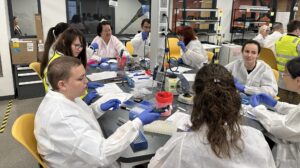 Group of people in a laboratory setting wearing white protective gowns and blue gloves, working with pipettes and lab equipment on a shared table.