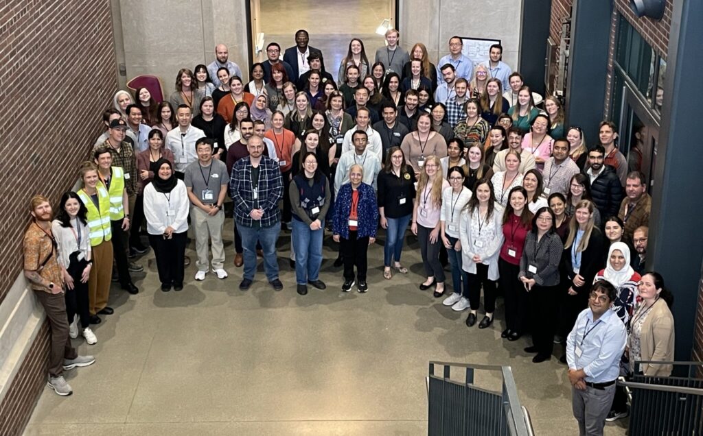 : Large group photo of event participants standing on outdoor steps in front of a building, gathered for a group picture during the symposium.