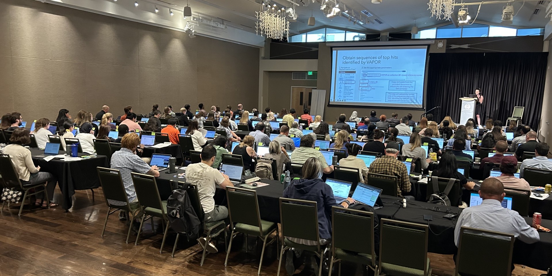 a group of members gathered in a room for a symposium, looking toward a stage with a screen and a presenter