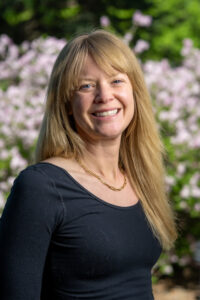 a person with long blonde hair smiles at the camera, with flowers in the background
