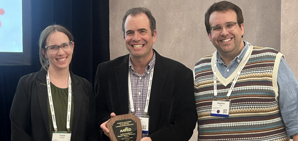 Three people smile at the camera, a person in the middle holding an award