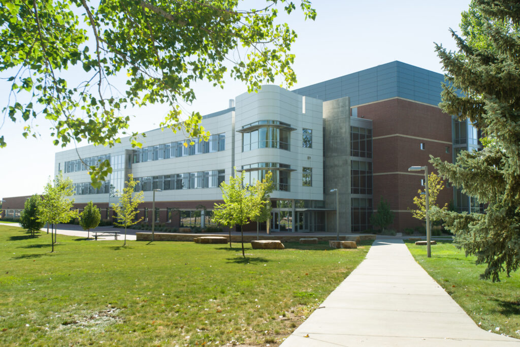 A modern multi‑story campus building with glass and metal accents is shown from a sidewalk bordered by grass and trees.
