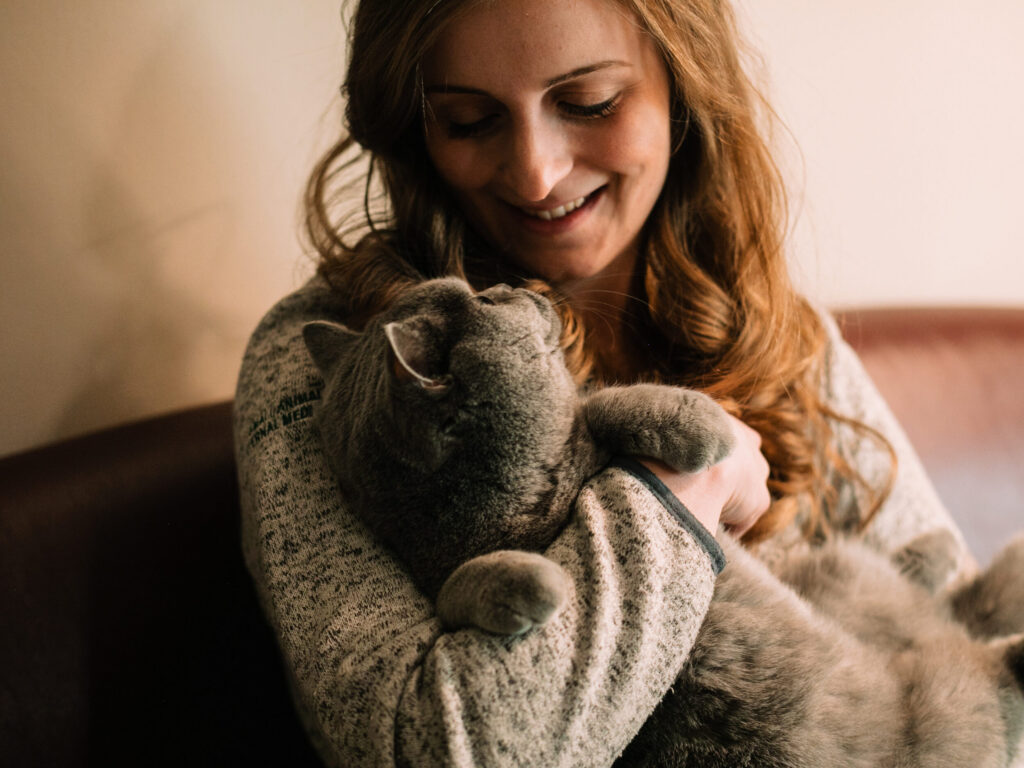 a person with wavy red hair holds a gray cat like a baby, looking down lovingly into the cat's eyes