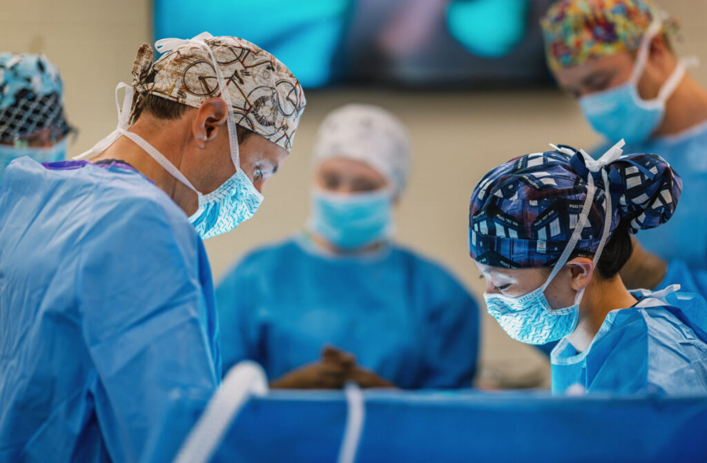 Several individuals wearing blue surgical gowns, disposable caps, and masks are gathered around an operating table in a brightly lit medical or veterinary surgical suite. They are focused on a procedure taking place out of view, with surgical instruments and sterile drapes visible in the foreground. A large monitor is mounted on the wall in the background.