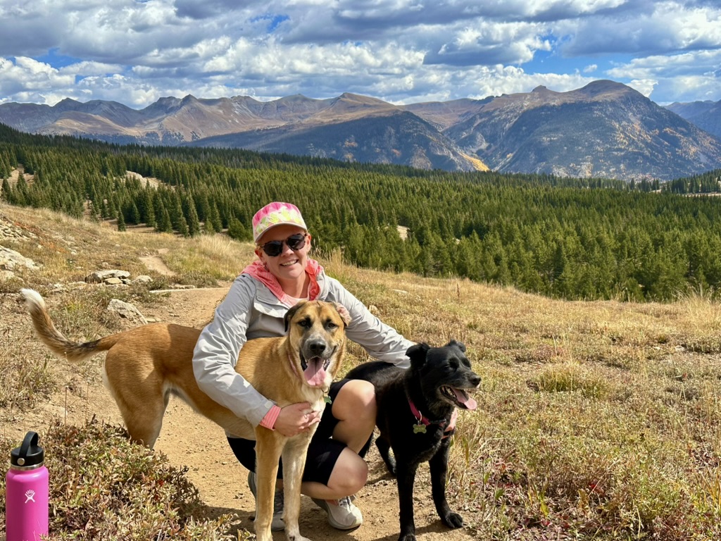 a person wearing a pink hat kneels with her her two dogs, one brown one black with a mountain landscape in the background